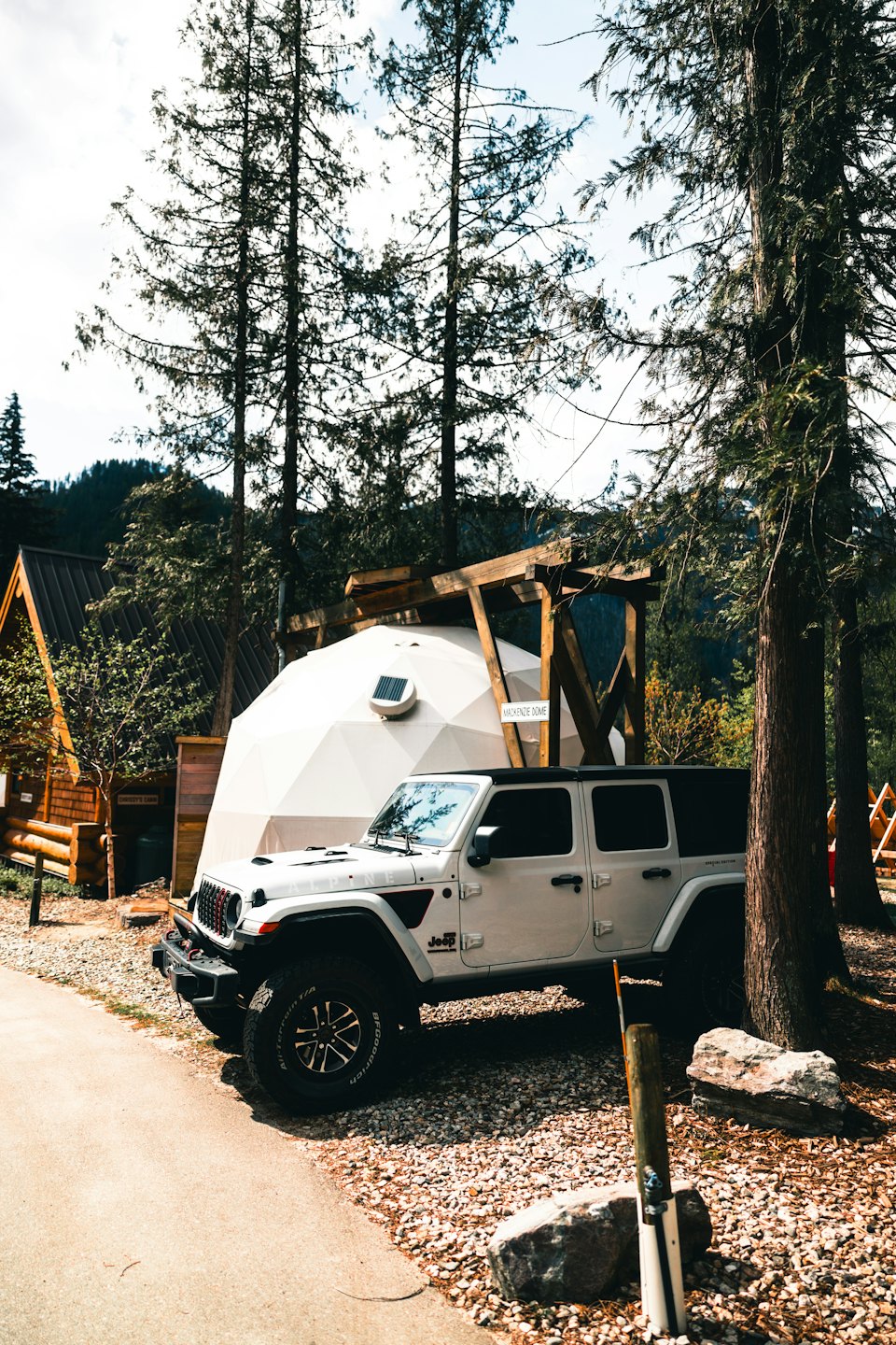 A white Jeep parked near a geodesic dome and wooden cabin.