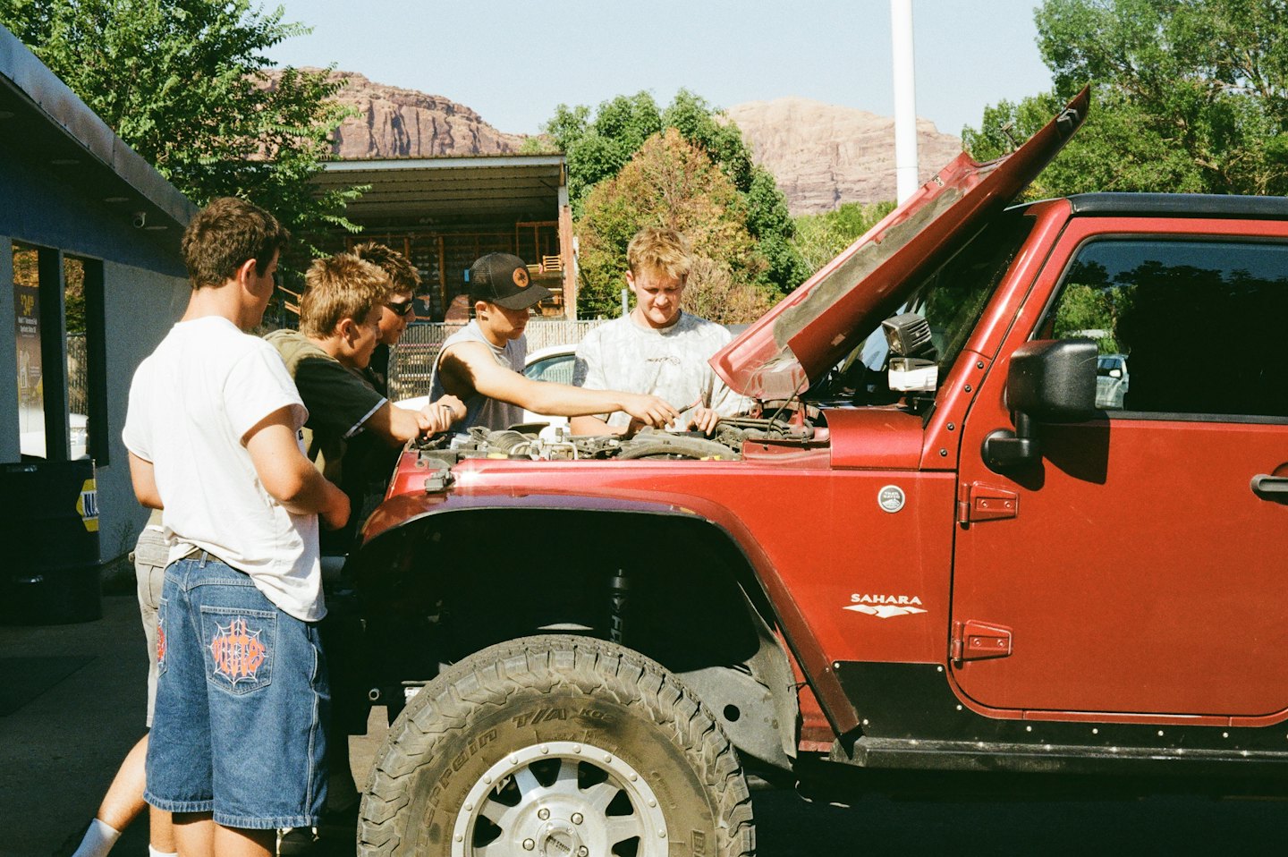 Four men inspect the engine of a red Jeep Wrangler.
